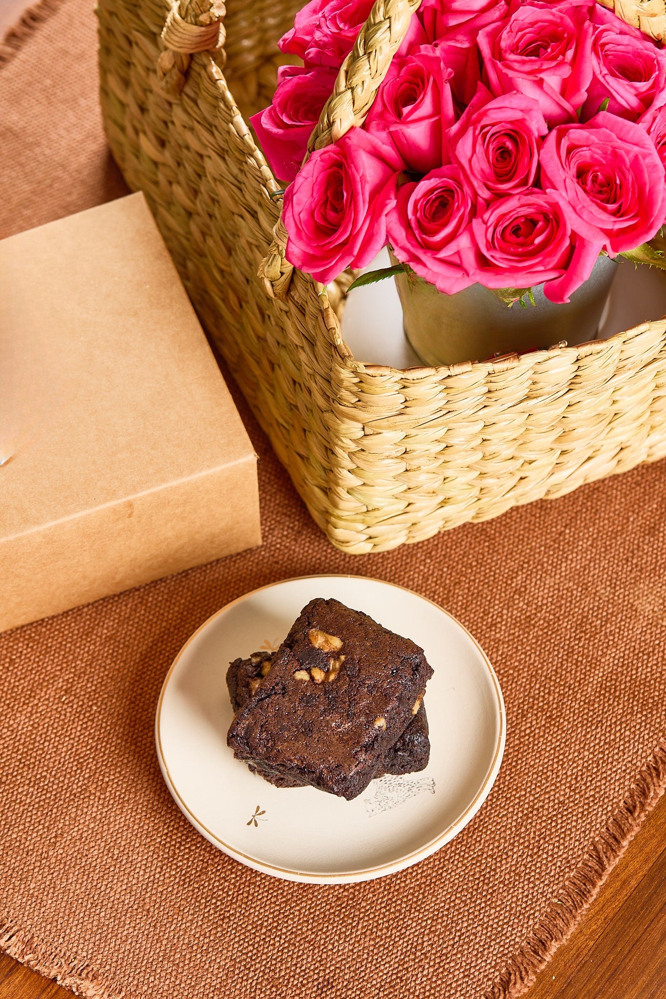 Brownie on a plate with a basket of pink roses and a gift box on a brown surface