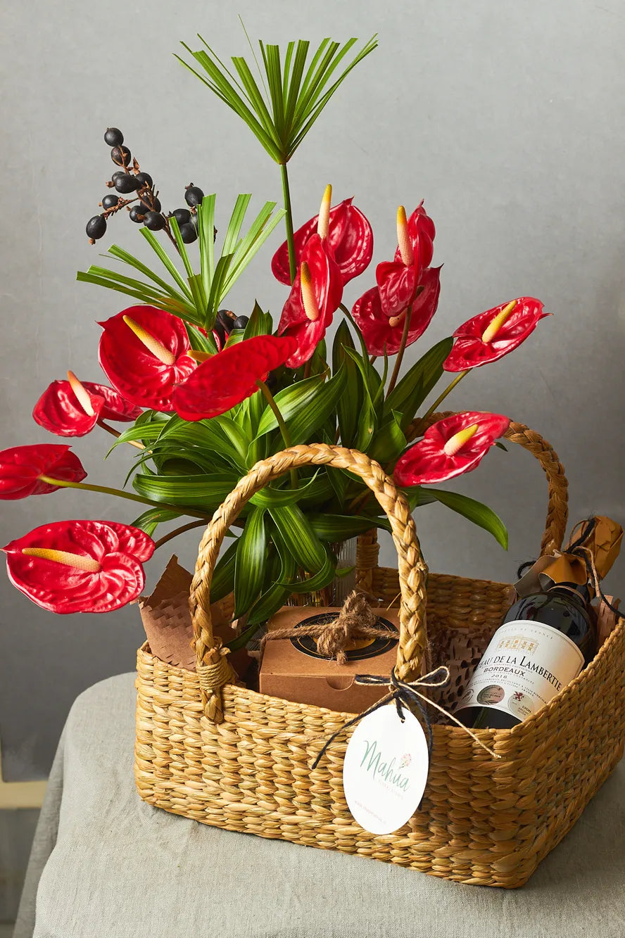 Water hyacinth basket with anthuriums  in a vase, a bottle of wine, and decorative items on a gray surface.