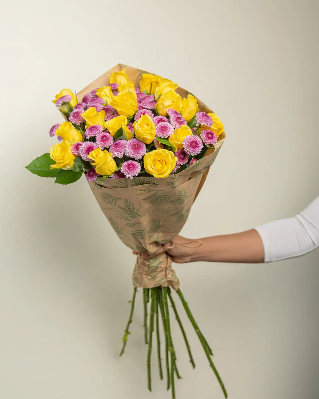 Bouquet of yellow and pink flowers wrapped in brown paper held by a person against a plain background