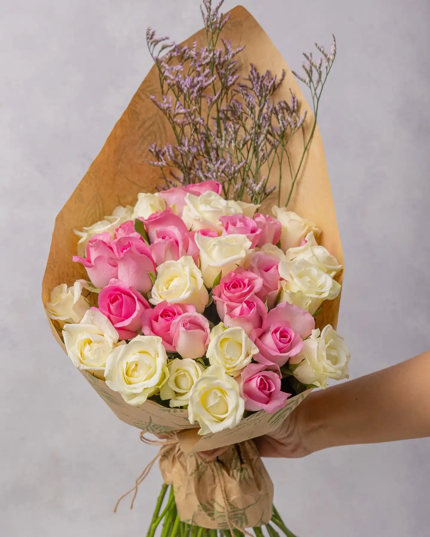 Bouquet of pink and white roses with lavender sprigs in a kraft wrap, held against a light gray background.